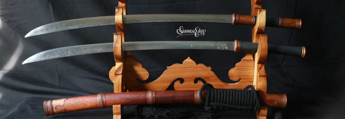Studio shot on black: two Burmese Dha blades on a carved wooden stand above a hardwood scabbard with copper fittings and black rope