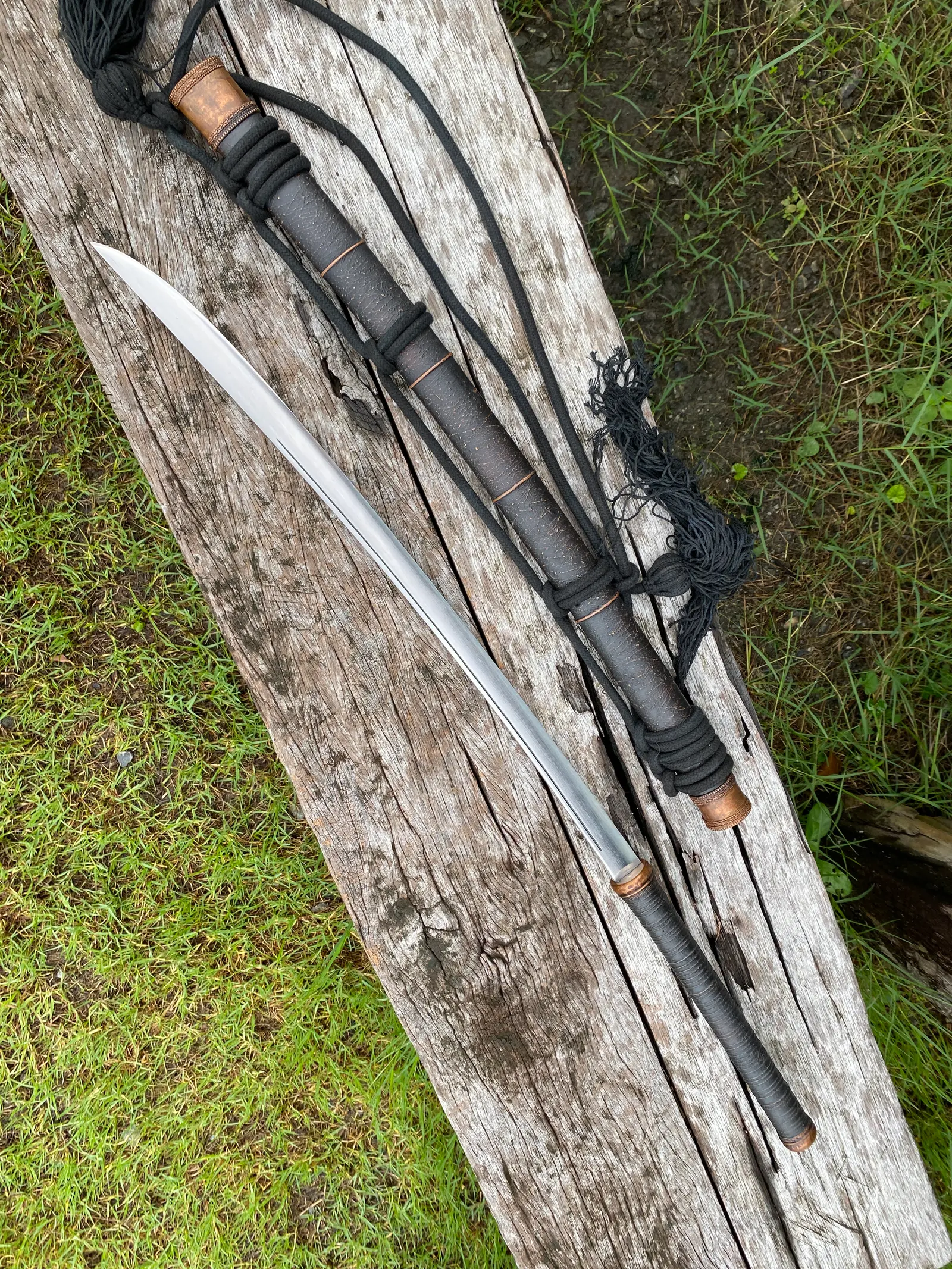 The sword drawn beside its black textured scabbard with copper bands and black carry rope, on a weathered plank