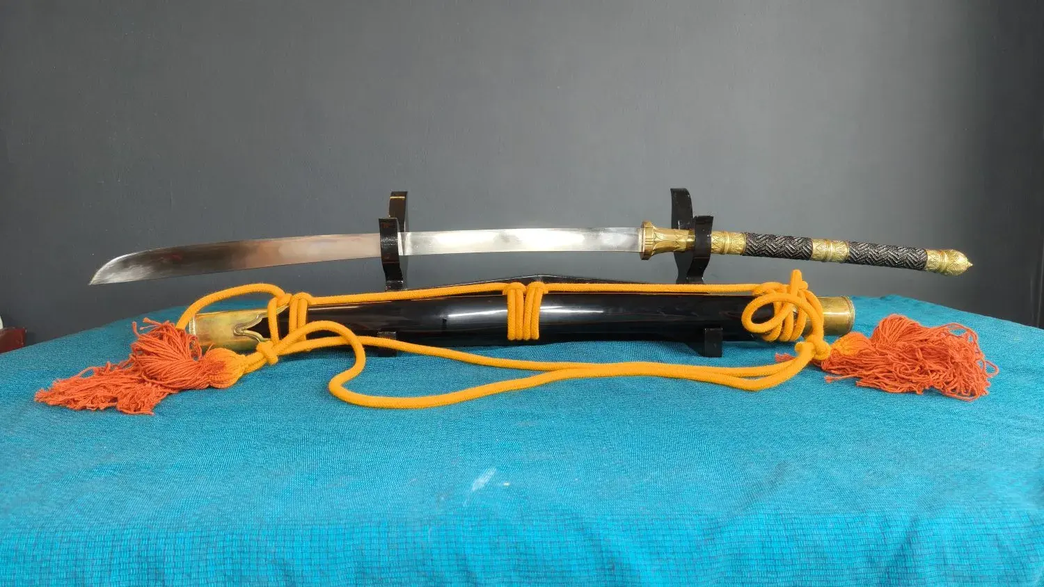 Side view on a black stand: drawn sword above the scabbard, orange rope coiled.