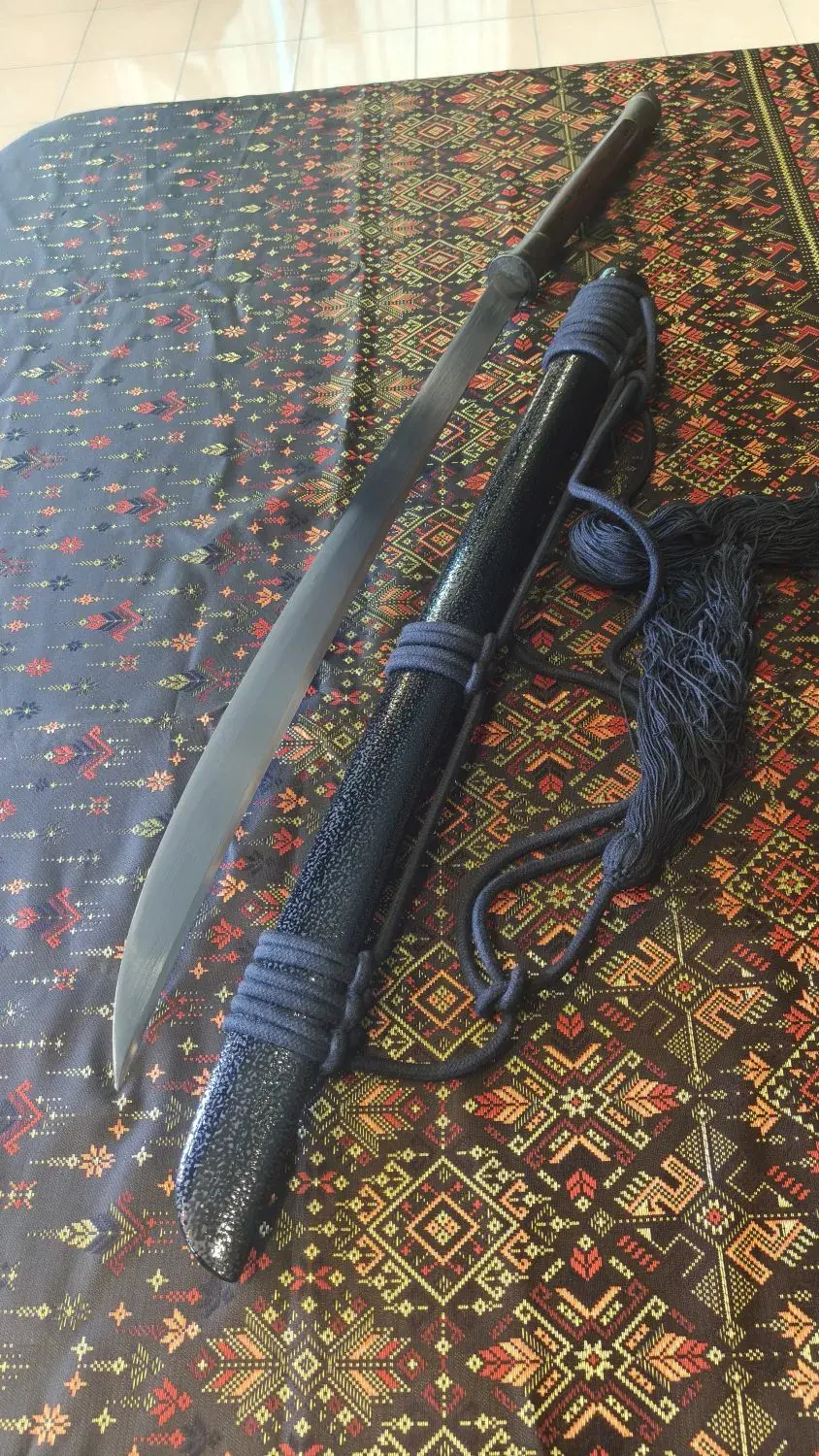 Burmese-Thai style piece seen from above on Thai woven cloth: blade beside a black textured scabbard with rope and tassels