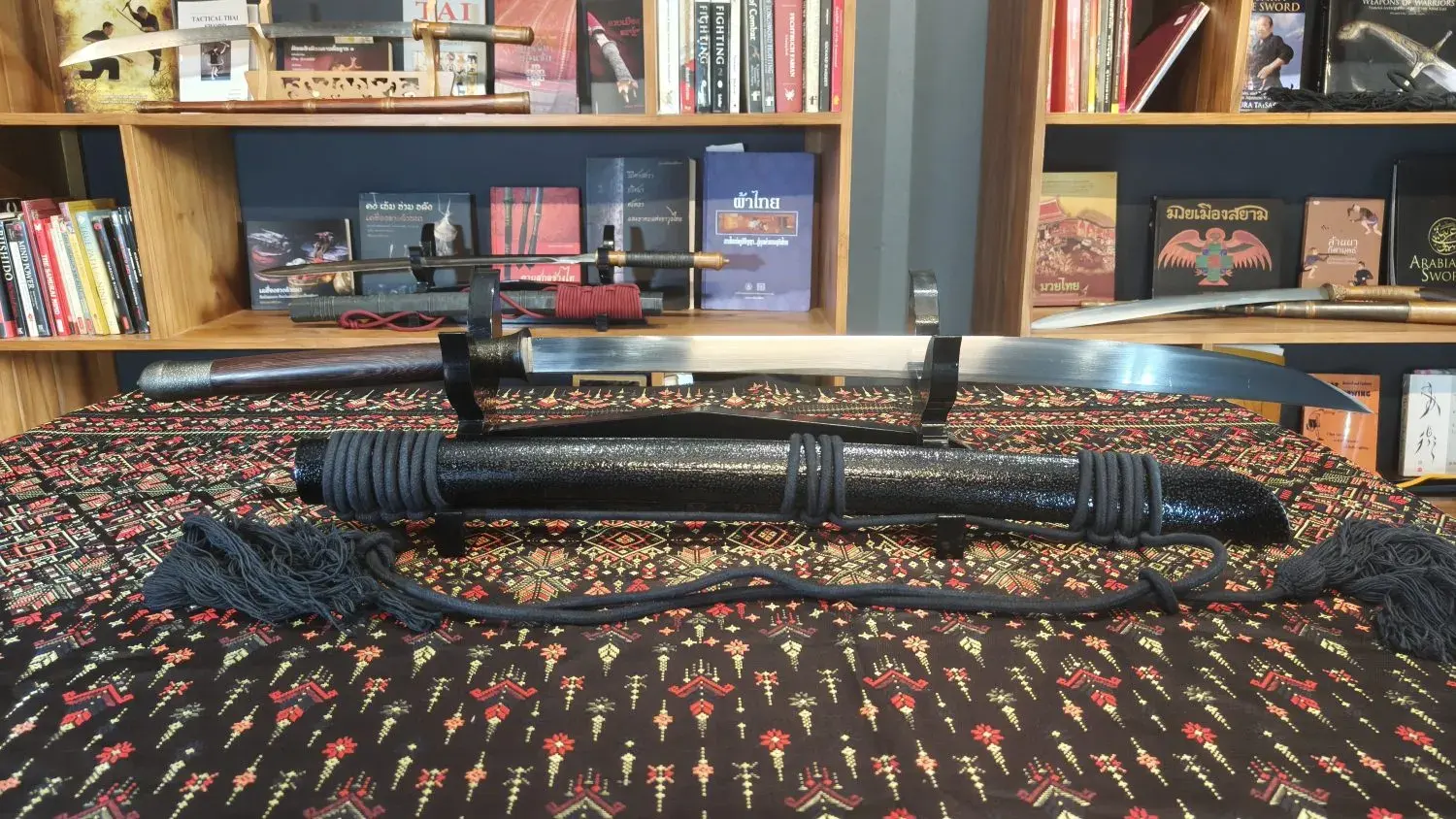 The ready-to-ship Burmese-Thai style piece on its stand in the showroom, drawn blade above a black textured scabbard