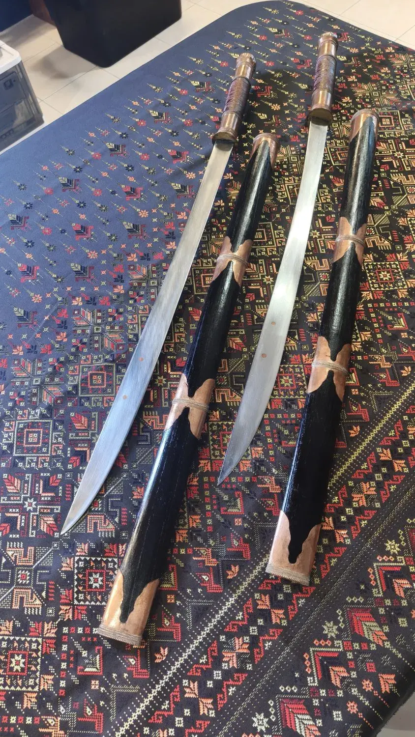 The Tai Dhaab set pair with copper fittings, drawn beside their black scabbards on Thai woven cloth