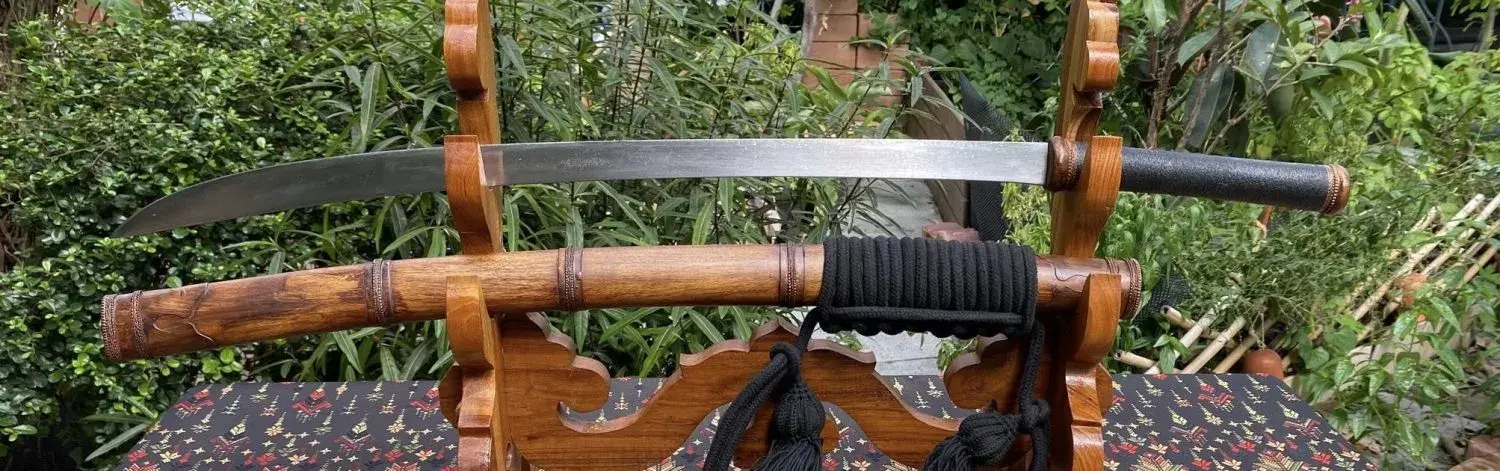 Basic Burmese Dha on a wooden stand in a garden: wooden scabbard with copper fittings and black rope